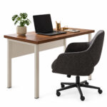 Mesa pé quadro com notebook, planta e materiais sobre o tampo, acompanhada de cadeira giratória estofada escura.