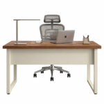 Mesa vista pelo lado externo, com painel central claro, notebook e luminária sobre o tampo e cadeira com apoio de cabeça atrás.