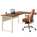 Mesa em L pé quadro em composição de trabalho Mesa em L com tampos amadeirados e pés metálicos claros, acompanhada de notebook, planta e cadeira giratória marrom.
