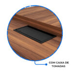 Detalhe da caixa de tomadas no tampo amadeirado Detalhe circular da tampa preta da caixa de tomadas embutida no tampo amadeirado, junto à borda elevada da mesa.