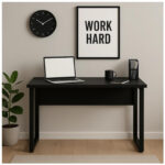 Mesa preta com pé quadro em home office com notebook Mesa preta com notebook, caneca e porta-canetas em ambiente com planta, relógio e quadro decorativo na parede.