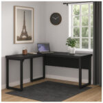 Mesa em L preta com notebook e planta em ambiente com janela, relógio, quadro decorativo e tapete escuro.