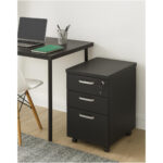 Gaveteiro preto ambientado ao lado de mesa de trabalho Gaveteiro preto de três gavetas ao lado de mesa com notebook e caderno verde, em ambiente com cadeira branca e tapete.
