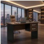 Mesa louro e preto com notebook em escritório ambientado Mesa louro e preto com duas gavetas, notebook e cadeira de encosto alto, diante de janelas com vista urbana.