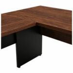Detalhe da junção dos tampos louro da mesa em L Detalhe do encontro dos tampos amadeirados da mesa em L, com veios aparentes e painel preto abaixo da junção.